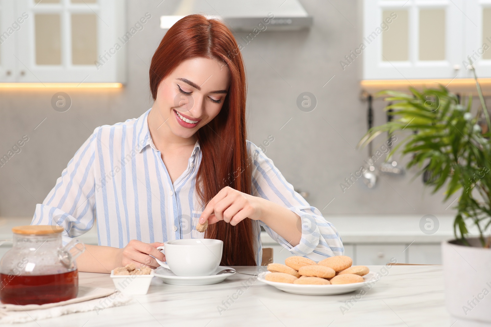 Photo of Happy woman with red dyed hair holding cookie above cup of tea at white marble table in kitchen, space for text