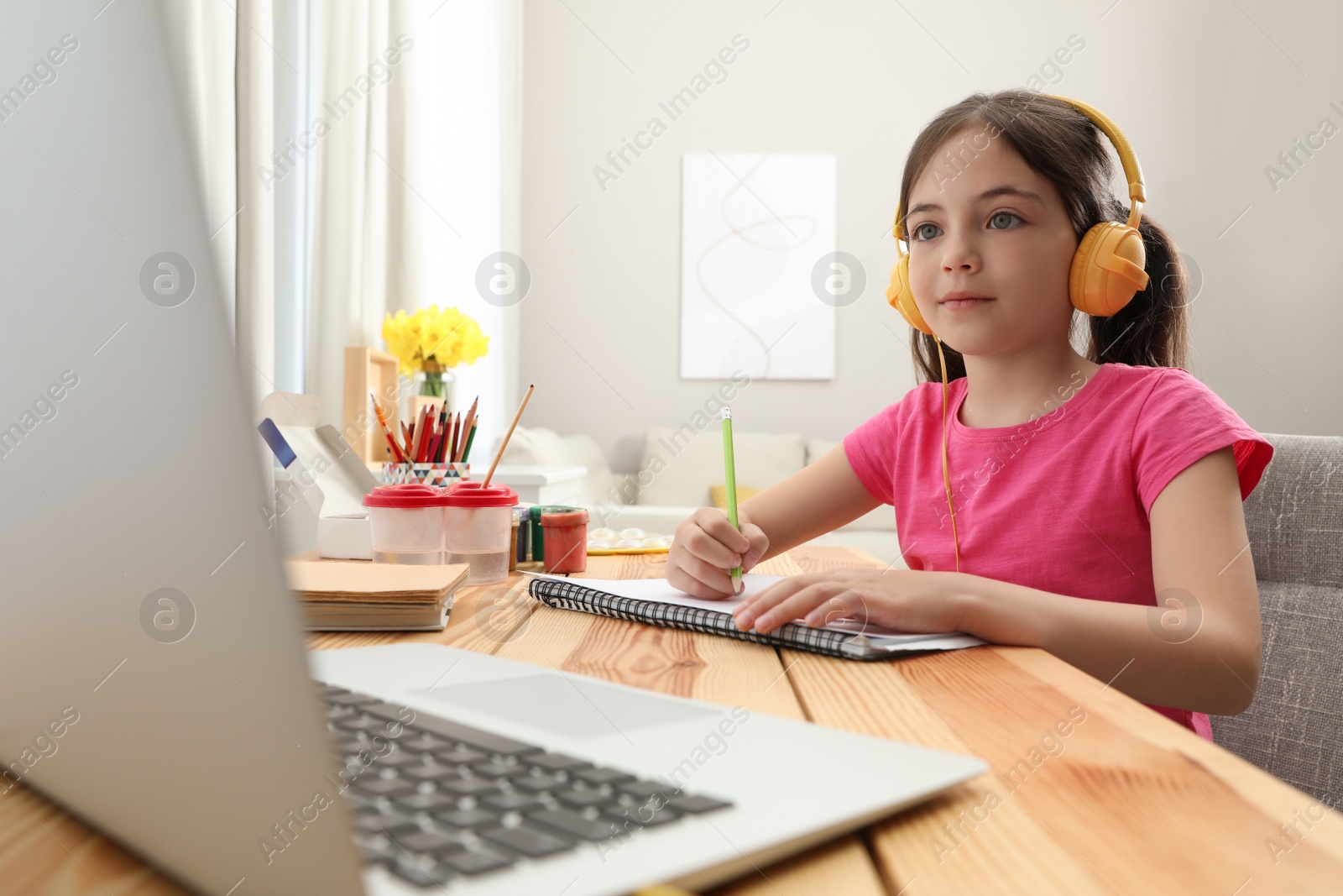 Photo of Little girl with headphones drawing on paper at online lesson indoors. Distance learning