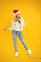 Photo of Emotional woman in Santa Claus hat singing with microphone on yellow background. Christmas music