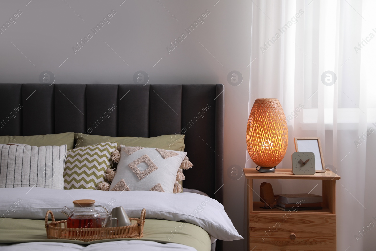 Photo of Wicker tray with teapot on comfortable bed, lamp and different decor on wooden bedside table in room. Stylish interior