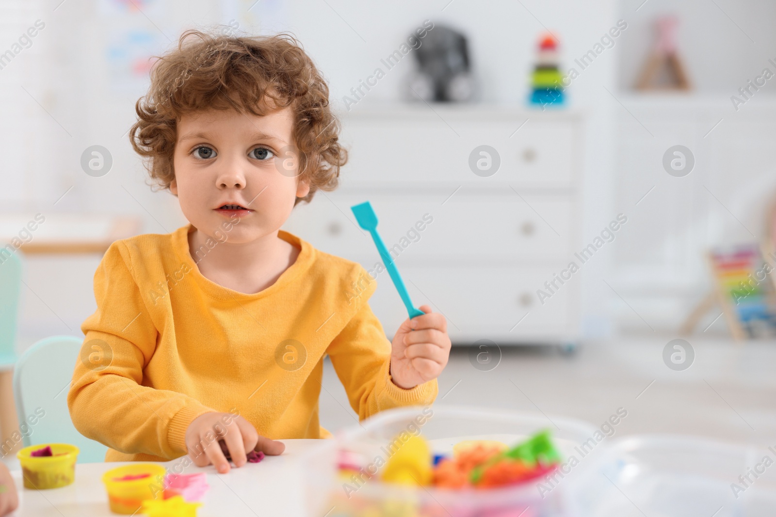 Photo of Cute little boy modeling from plasticine at white table in kindergarten. Space for text