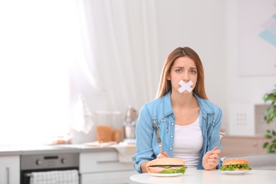 Emotional young woman with taped mouth and burgers at table in kitchen. Healthy diet