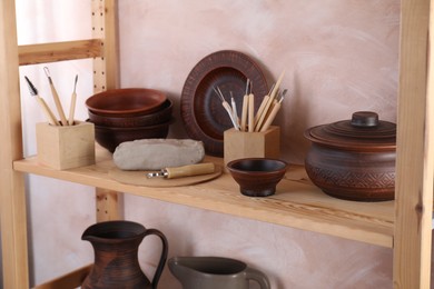 Photo of Set of different crafting tools and clay dishes on wooden rack in workshop