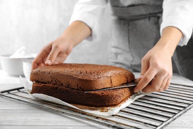 Photo of Woman cutting homemade chocolate cake into layers at white wooden table, closeup