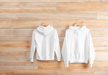 Photo of New hoodie sweaters with hangers on wooden wall. Mockup for design