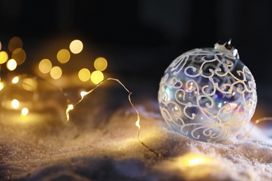 Photo of Christmas ball and fairy lights on white snow outdoors. Space for text
