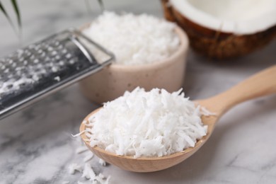 Photo of Coconut flakes in wooden spoon and grater on white marble table, closeup