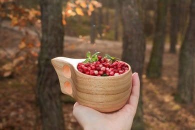 Photo of Woman holding wooden cup with tasty lingonberries in forest, closeup