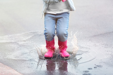 Photo of Little girl jumping in rubber boots under rain on street, closeup