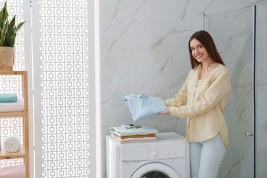 Happy woman with clean clothes near washing machine in bathroom. Laundry day