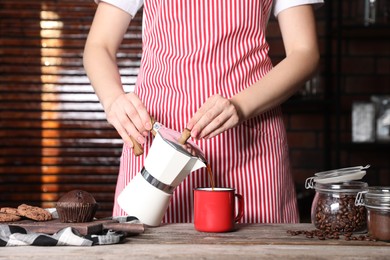 Photo of Woman pouring aromatic coffee from moka pot into cup at wooden table indoors, closeup