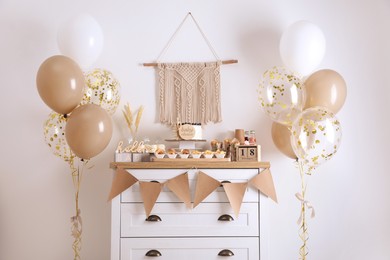 Photo of Baby shower party. Different delicious treats on white wooden chest of drawers and decor near light wall