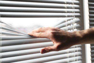 Photo of Man separating slats of white blinds indoors, closeup