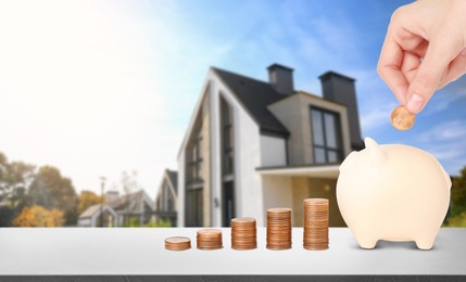 Image of Woman putting coin into piggy bank at stone table and blurred view of beautiful house. Mortgage concept