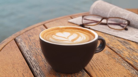 Photo of Cup of delicious coffee, eyeglasses and newspaper on wooden table, closeup