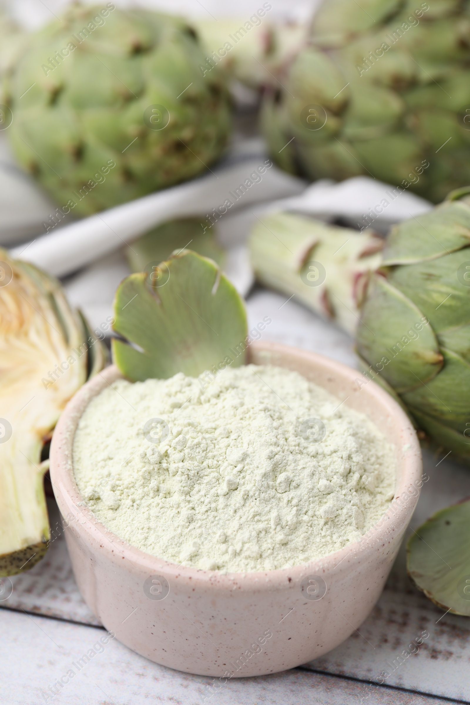Photo of Bowl with powder and fresh artichokes on white wooden table, closeup