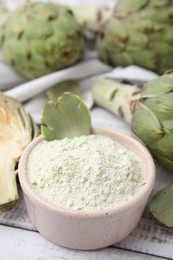 Photo of Bowl with powder and fresh artichokes on white wooden table, closeup