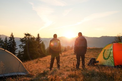 Men near camping tents in mountains at sunset, back view
