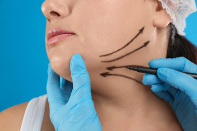 Doctor drawing marks on woman's face for cosmetic surgery operation against blue background, closeup. Double chin problem