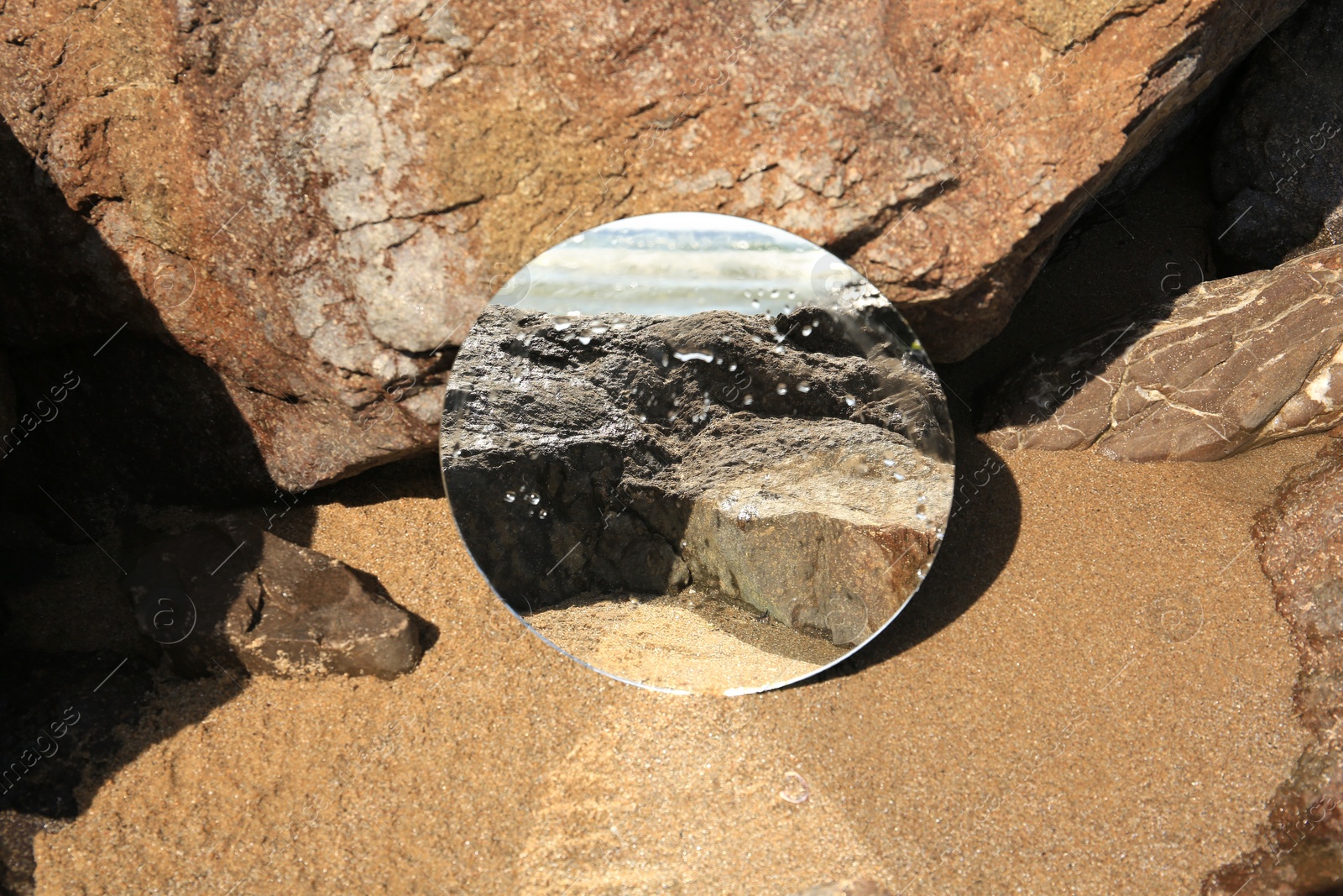 Photo of Round mirror reflecting seashore on sand near stones outdoors