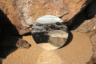 Photo of Round mirror reflecting seashore on sand near stones outdoors
