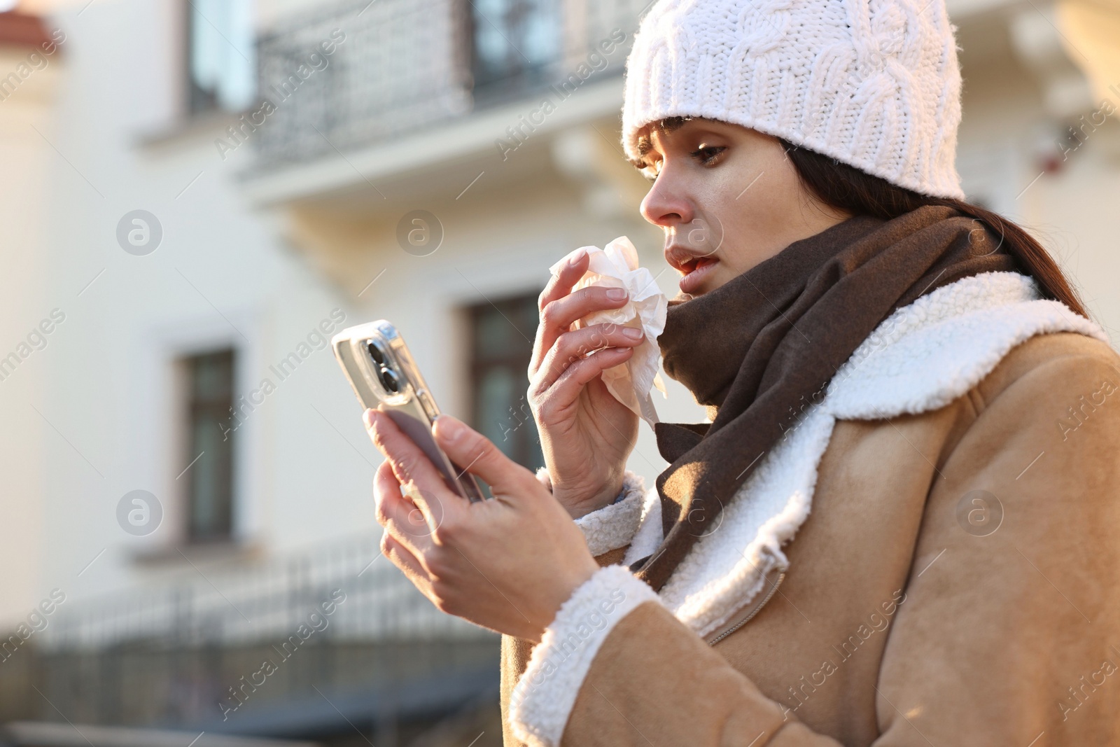 Photo of Woman with tissue blowing runny nose while using phone outdoors. Cold symptom