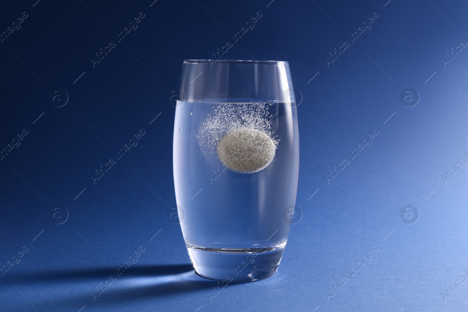 Photo of Effervescent pill dissolving in glass of water on blue background