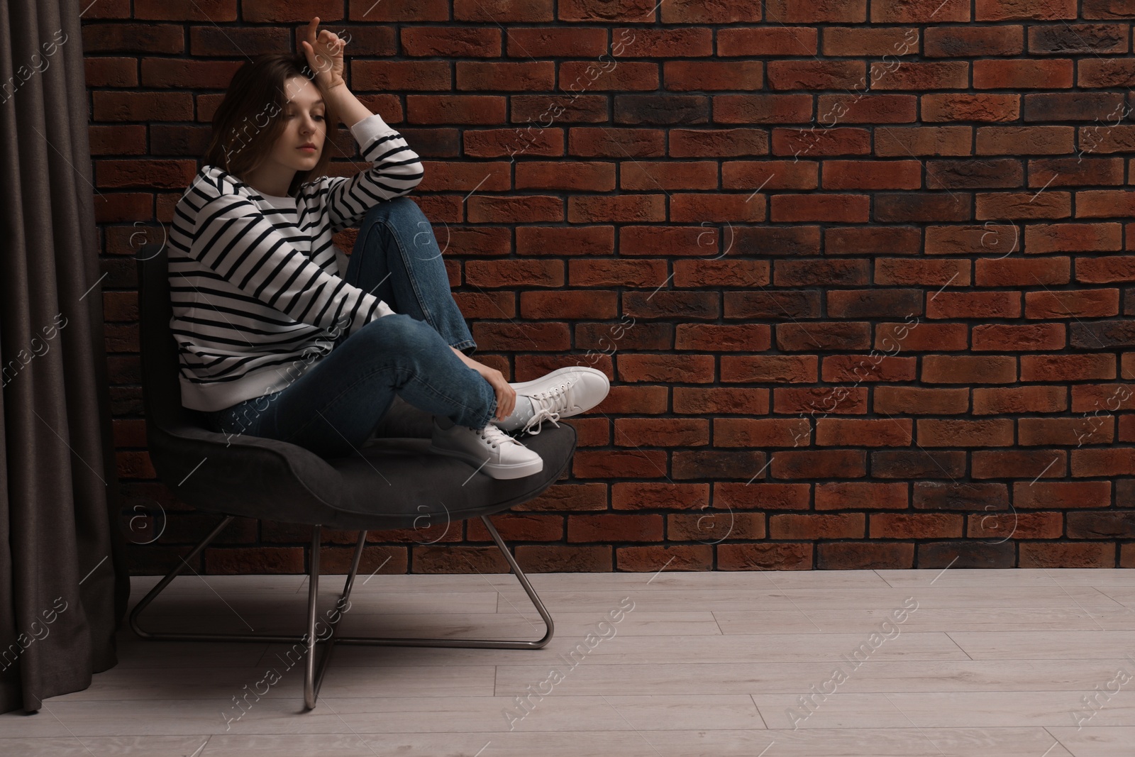 Photo of Sad young woman sitting on chair near brick wall indoors, space for text