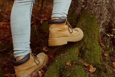 Photo of Woman in stylish boots standing near tree roots on ground covered with autumn leaves in forest