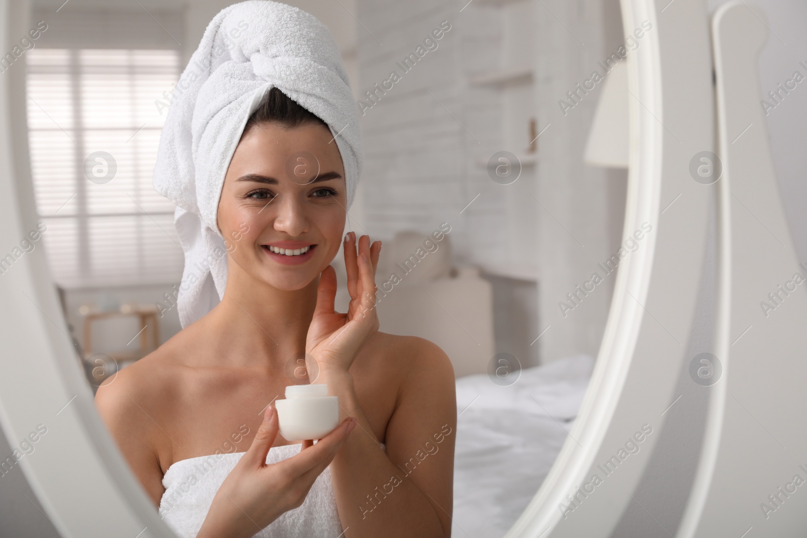 Photo of Beautiful young woman with hair wrapped in towel applying cream near mirror indoors