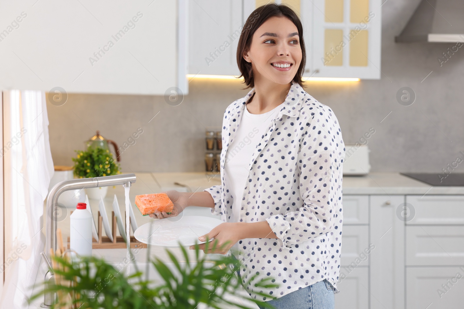 Photo of Happy young woman washing plate above sink in modern kitchen