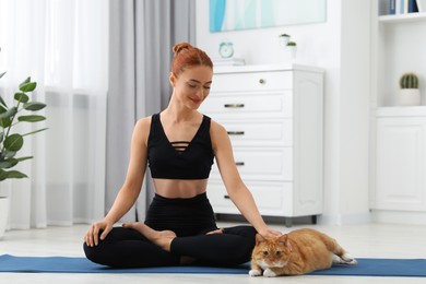 Beautiful woman petting cute red cat on mat at home. Practicing yoga