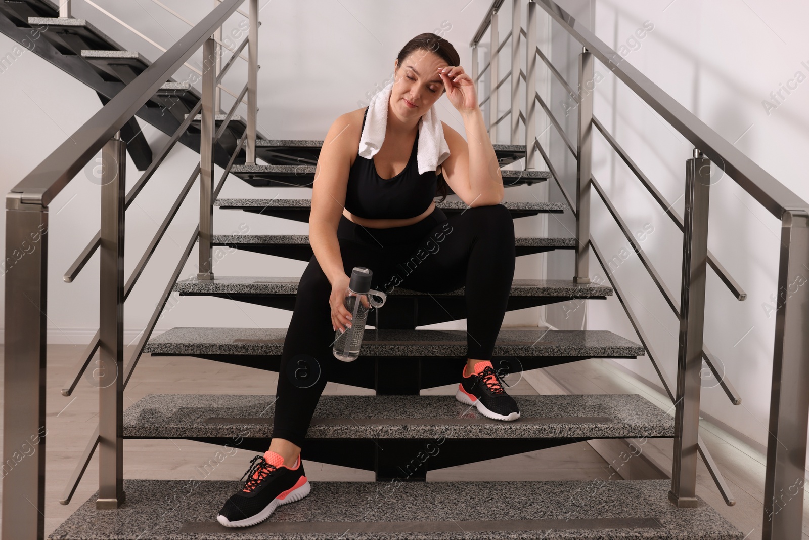 Photo of Tired overweight woman with towel and bottle of water sitting on stairs indoors