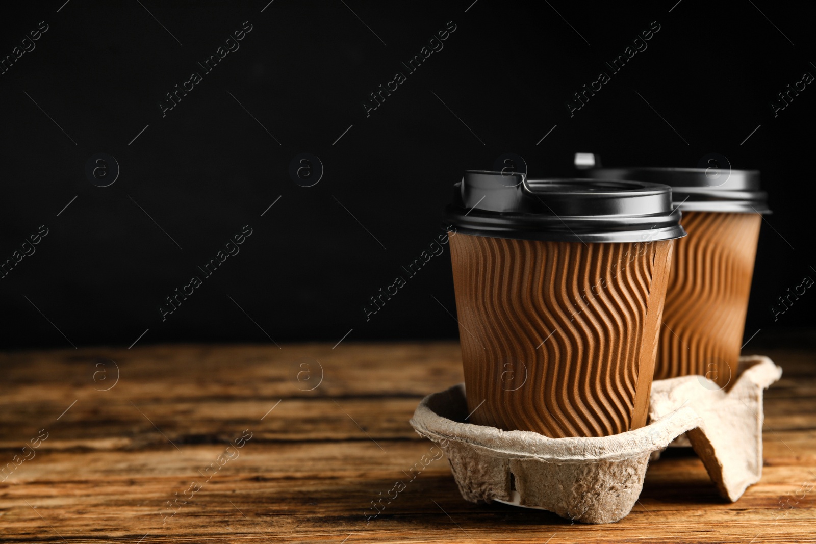 Photo of Takeaway paper coffee cups in cardboard holder on wooden table against black background, space for text