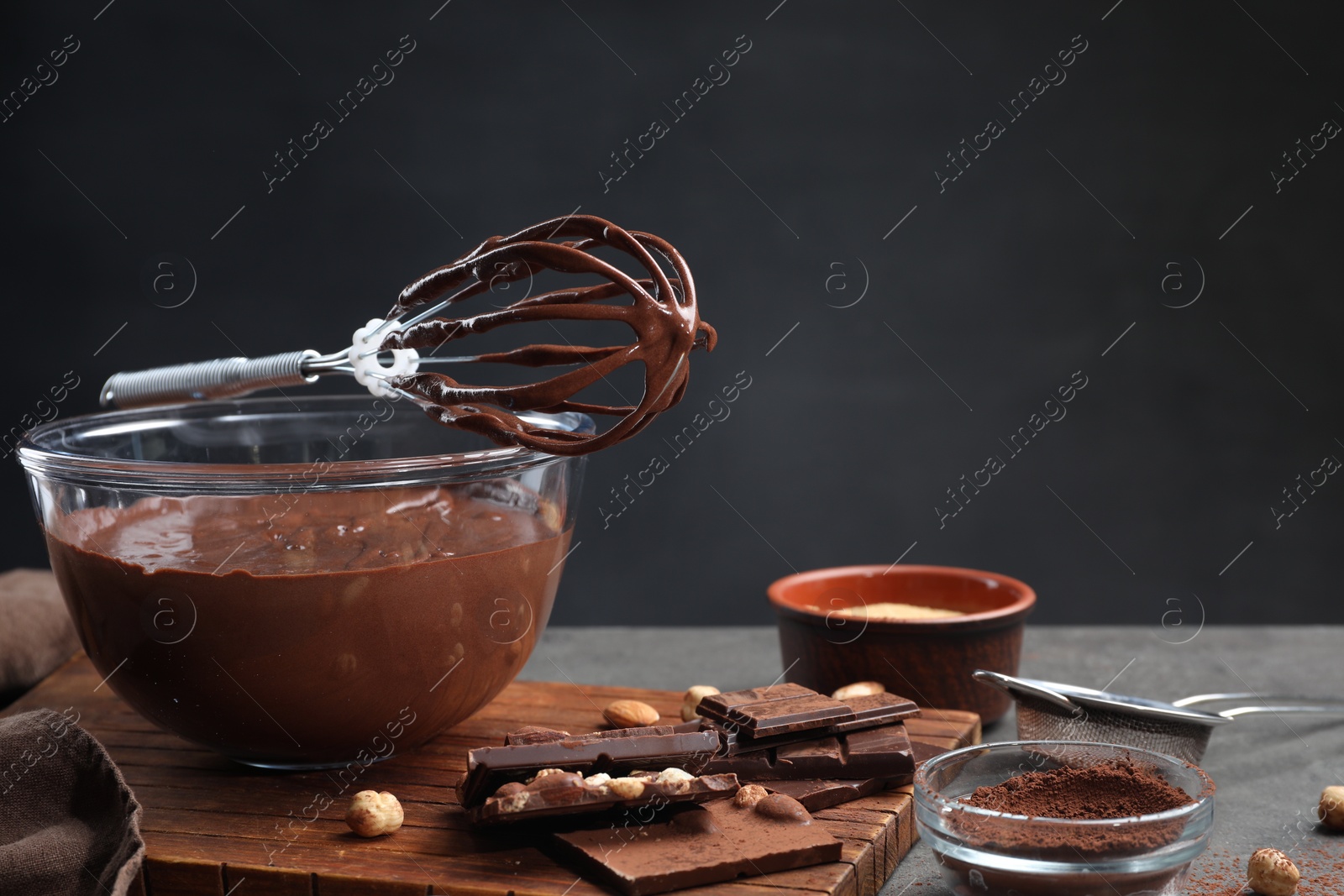 Photo of Bowl of chocolate cream, whisk and ingredients on gray table against dark background, space for text