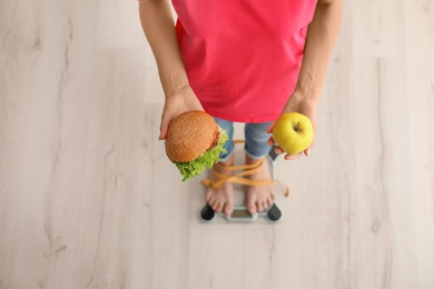 Photo of Woman holding tasty sandwich and fresh apple while measuring her weight on floor scales, top view. Choice between diet and unhealthy food