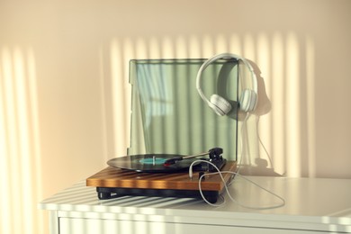 Photo of Stylish turntable with vinyl disc and headphones on white chest of drawers at home