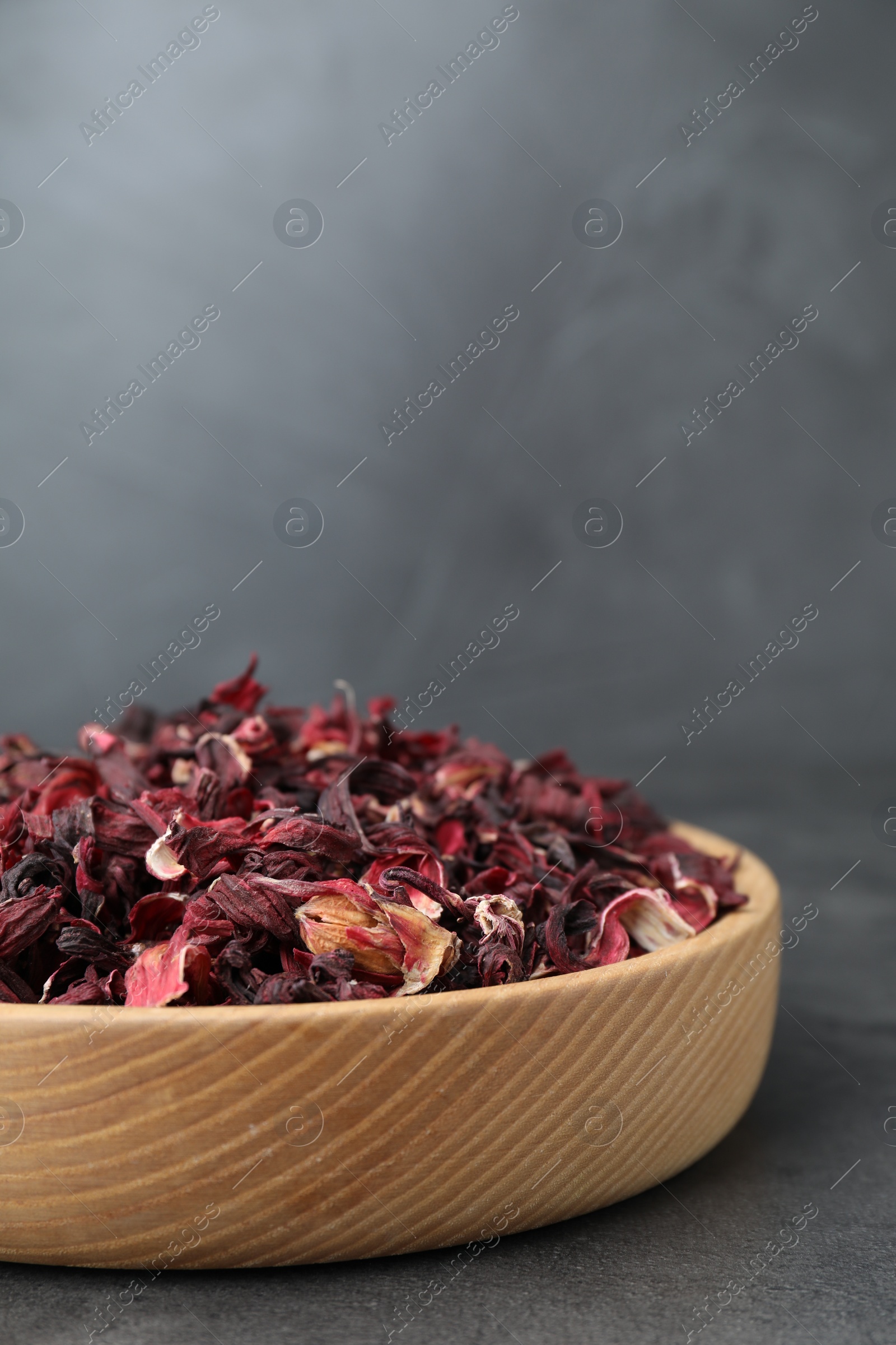 Photo of Hibiscus tea. Wooden bowl with dried roselle calyces on grey table, closeup. Space for text