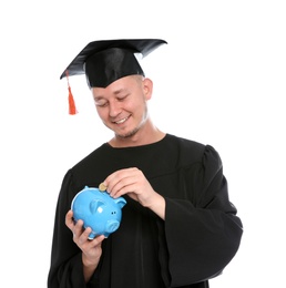 Portrait of young graduate putting coin into piggy bank on white background