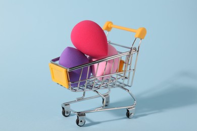 Photo of Makeup sponges in small shopping cart on light blue background, closeup