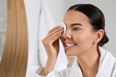 Photo of Beautiful woman removing makeup with cotton pad near mirror indoors