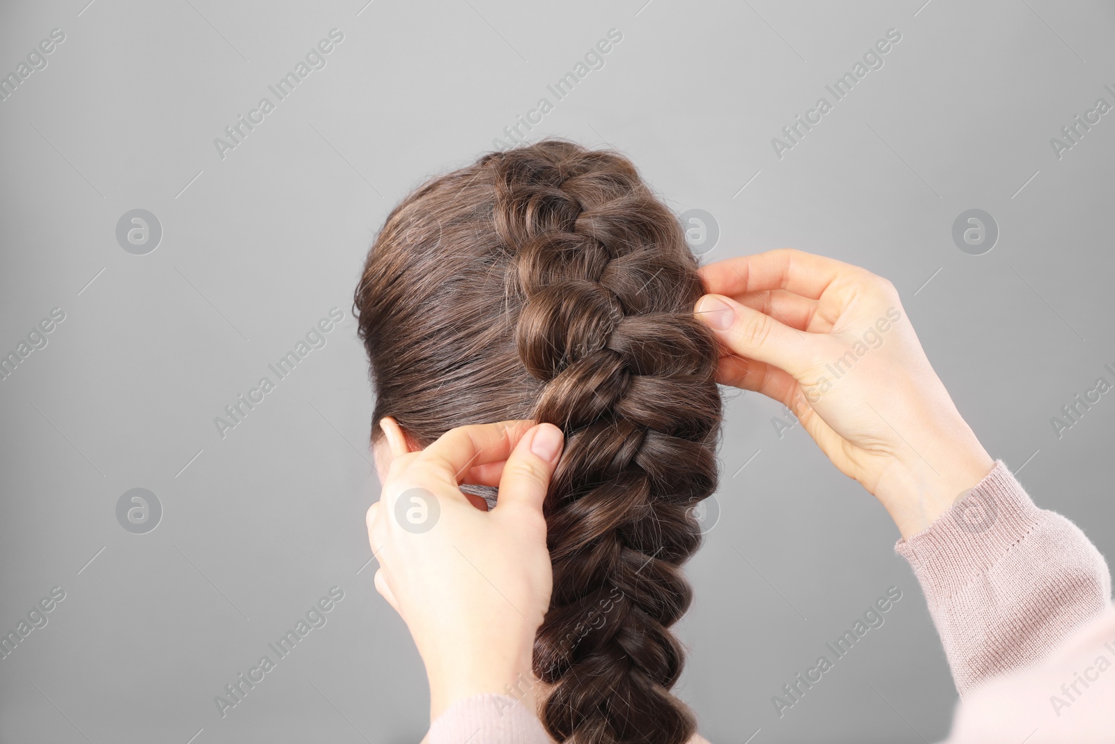 Photo of Professional stylist braiding woman's hair on grey background, closeup