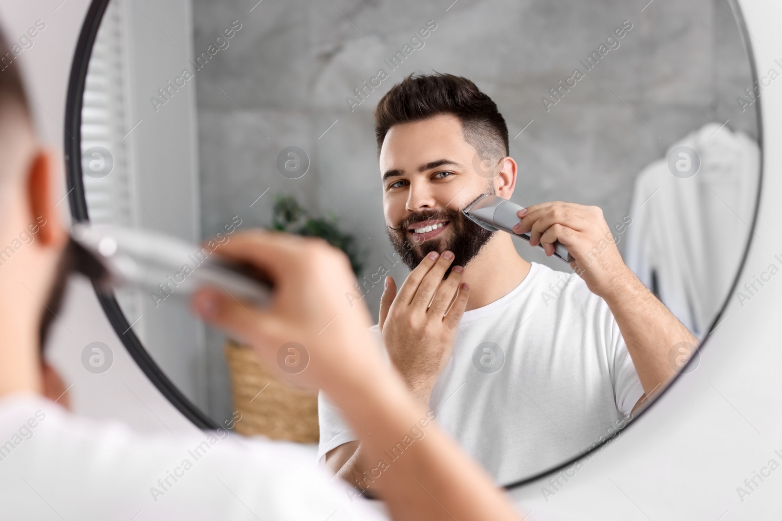 Photo of Handsome young man trimming beard near mirror in bathroom