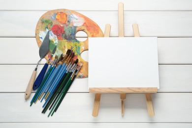Photo of Easel with blank canvas, brushes and palette on white wooden table, flat lay. Space for text