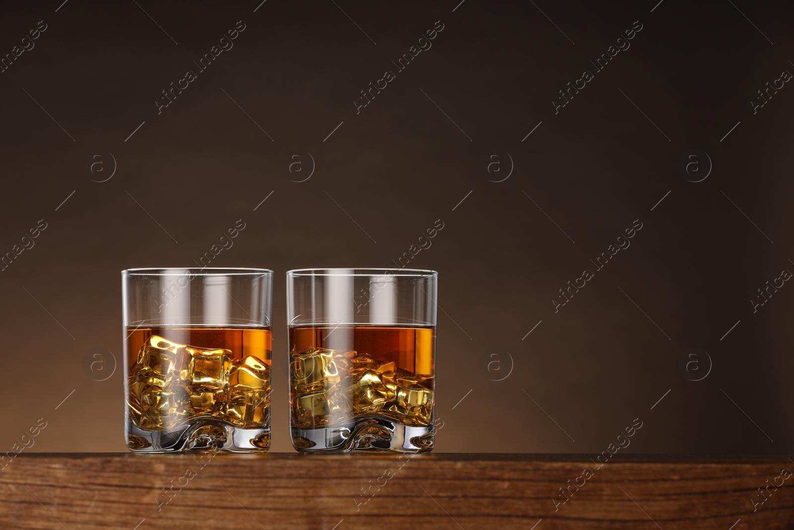 Photo of Whiskey with ice cubes in glasses on wooden table against brown background, space for text
