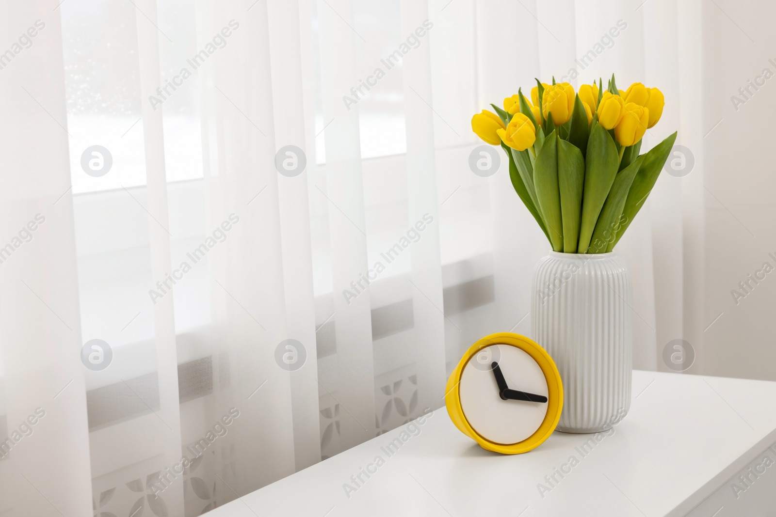 Photo of Spring interior. Beautiful yellow tulips and clock on white chest of drawers near window, space for text