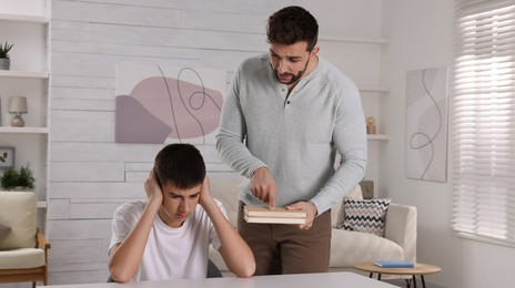 Photo of Strict father with books scolding his son at home. Teenager problems