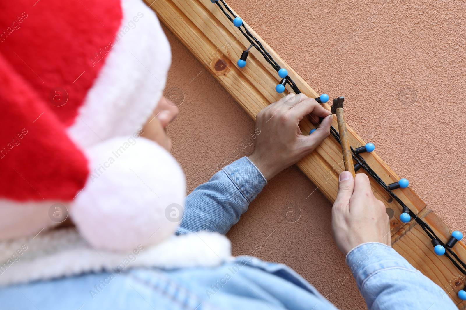 Photo of Man in Santa hat decorating house with Christmas lights outdoors, back view