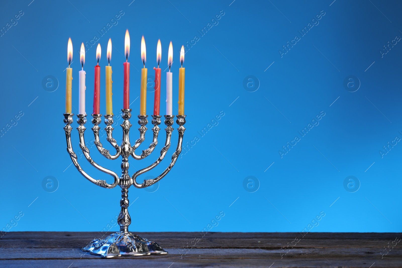 Photo of Hanukkah celebration. Menorah with burning candles on wooden table against blue background, space for text