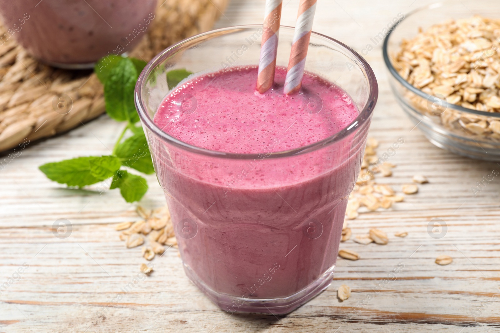 Photo of Glass of blackberry smoothie with straws, mint and oatmeal on light wooden table, closeup